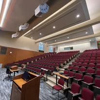 A modern lecture hall featuring tiered seating with burgundy chairs, a wooden podium, and overhead lighting, designed for educational presentations.