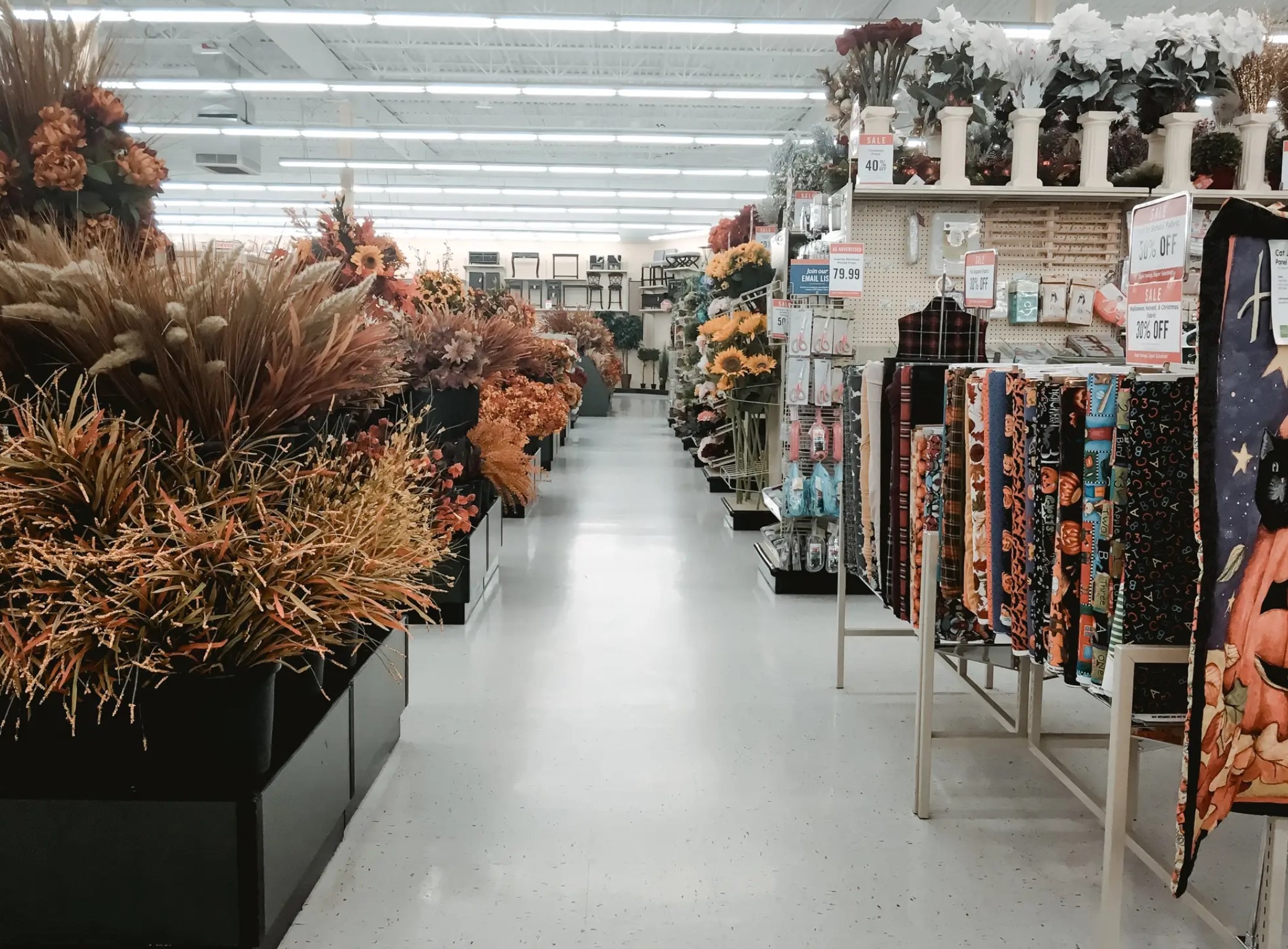 A vibrant aisle in a craft store showcases seasonal decorations, featuring artificial flowers in autumn hues and colorful fabrics with Halloween themes.